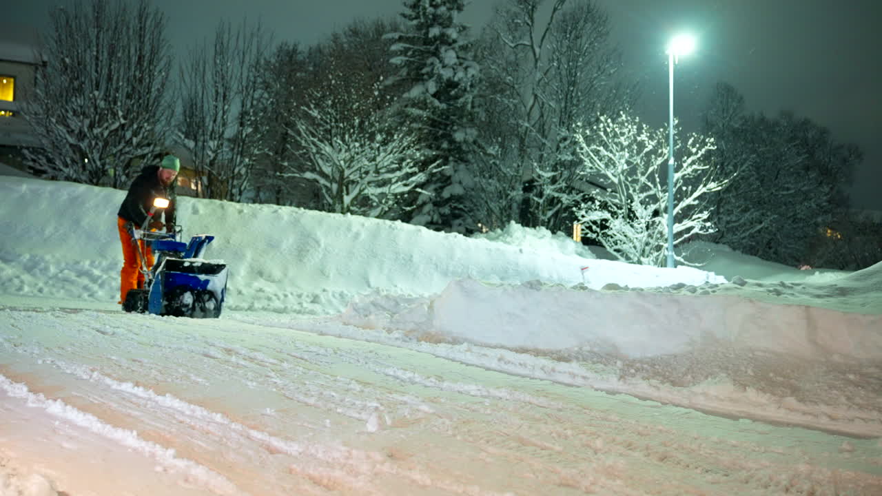 A man using snowblower gasoline machine to clear deep snow from front of residential house after heavy snowfall at night