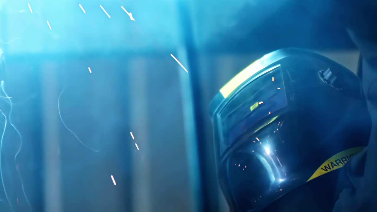 Primo piano di un saldatore che indossa un casco da saldatura durante la lavorazione dei metalli, circondato da un bagliore blu e da brillanti scintille volanti. Scena di un ambiente di lavoro industriale incentrata su sicurezza, precisione e maestria tecnica.