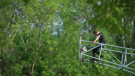 a young boy sits on blue iron outdoor equipment with his head bowed, while a soccer ball is visibly stuck in the iron framework, with trees in the background