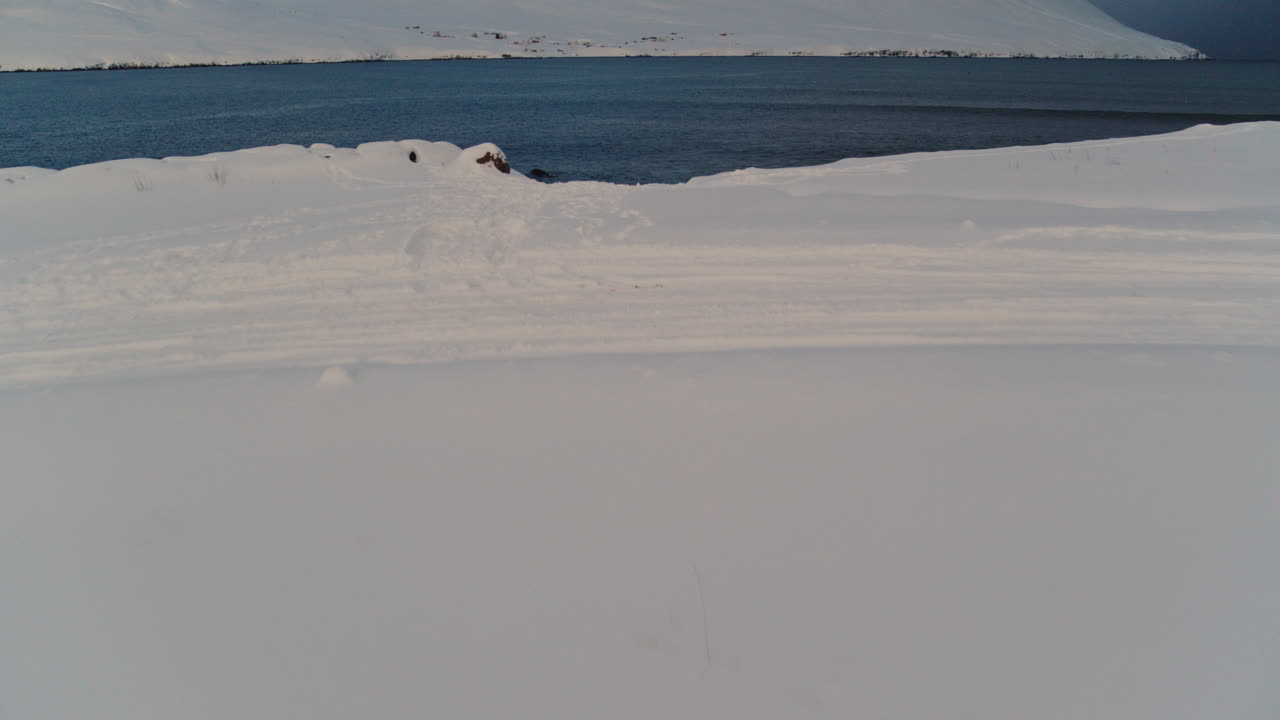 Tranquil Icelandic coast with snow and waves reflecting soft light under Arctic winter skies, drone descend reveal grand land and seascape