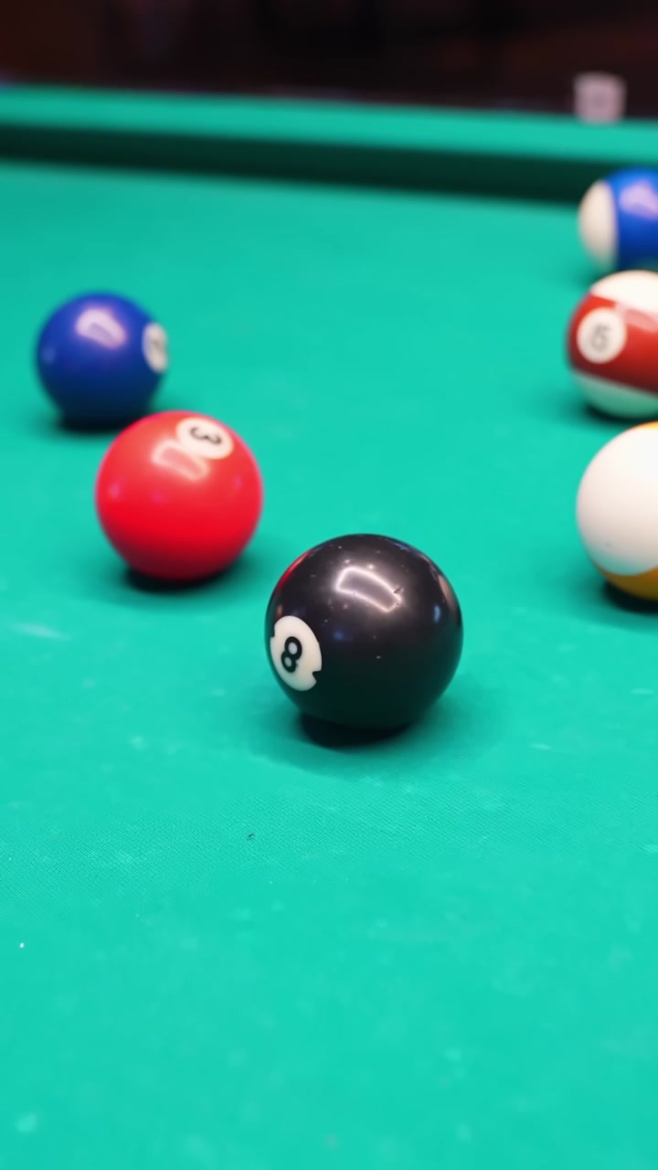 Vertical close-up of black 8 ball on green felt with colorful billiard balls blurred in the background, shallow focus for sports bar, competition, and game night b-roll.