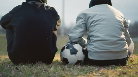 close-up of two people sitting on a grassy field, with one adjusting his hand on a soccer ball, during twilight, with blurred background lights