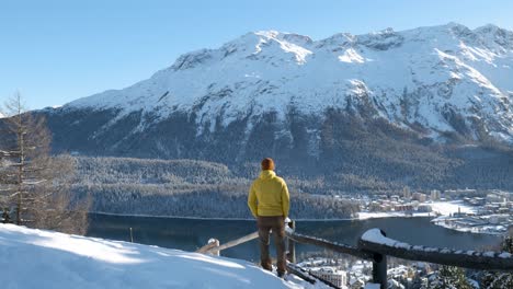 Kaukasischer-Junger-Mann-Mit-Gelber-Jacke-Und-Mütze,-Der-An-Einem-Sonnigen-Wintertag-In-St.-Den-Blick-Auf-Einen-See,-Eine-Stadt-Und-Berge-Genießt