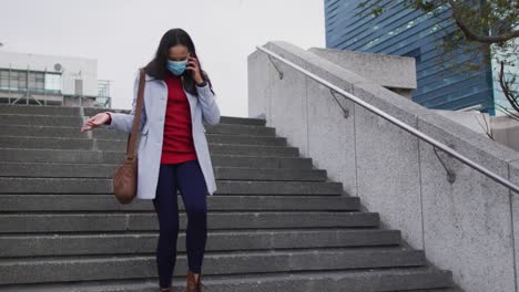 asian woman wearing face mask walking down the stairs and talking on smartphone