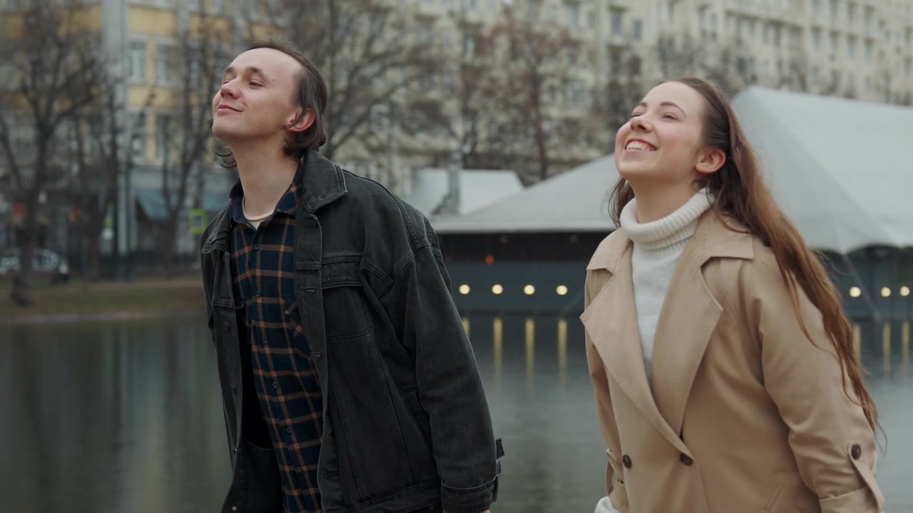 Two people man and woman engage in conversation by calm water body with buildings and trees. scene features soft overcast lighting natural outdoor setting details including bare branches
