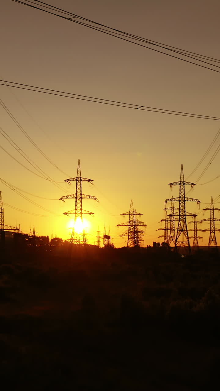 Dark silhouette of transmission towers at sunset. High-voltage electric lines against the orange setting sun. Electricity distribution. Vertical video