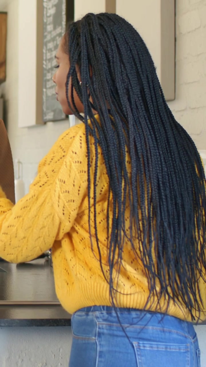 Donna appoggiata al bancone della cucina, con maglione giallo e jeans blu, che si sistema i capelli, indossando un grembiule.