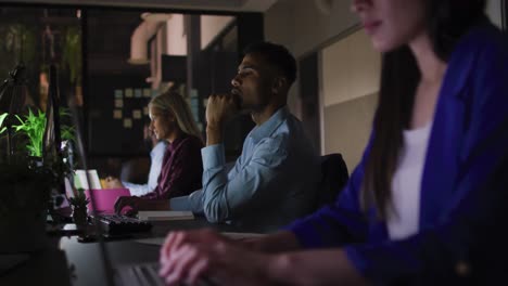 video of four diverse male and female colleagues sitting at desks using computers at night in office