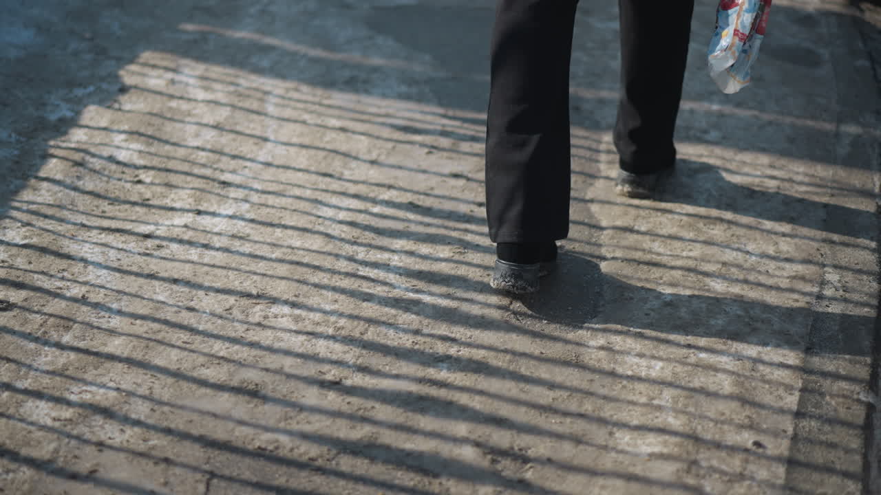low view legs in boots walking across pavement covered with striped shadows from fence under bright winter sun, gritty concrete, denim jeans and long coat moving forward during cold urban commute