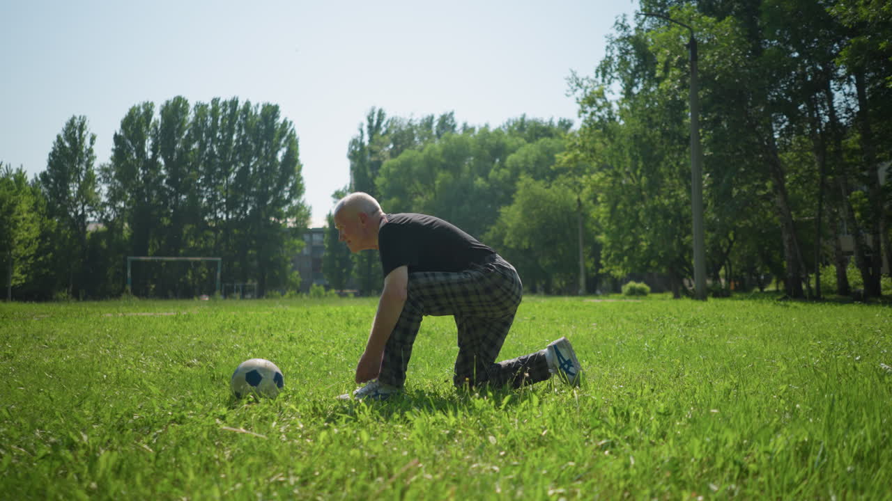 초록색 에서 한 다리에 무을 고 신중하게 신발을 매고 있는 노인, 그 에 축구공이 놓여 있다.