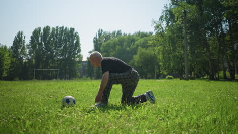 a side view of an elderly man kneeling on one leg in a green field, carefully tying his shoelace, a soccer ball lies close by him