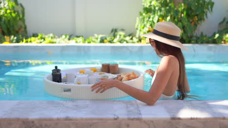 Slim-woman-standing-in-the-swimming-pool-and-touching-a-floating-breakfast,-a-bali-style-meal-which-is-a-popular-tourist-attraction