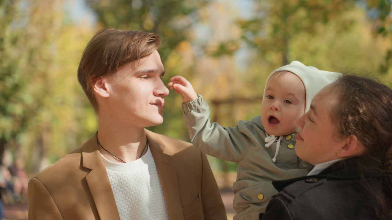 Unge forældre holder baby i parken, solbeskinnet efterårsscene med gyldne blade, far smiler, mens mor vugger spædbarn, blid berøring og legende interaktion, oprigtigt livsstilsportræt, der fanger varmen.
