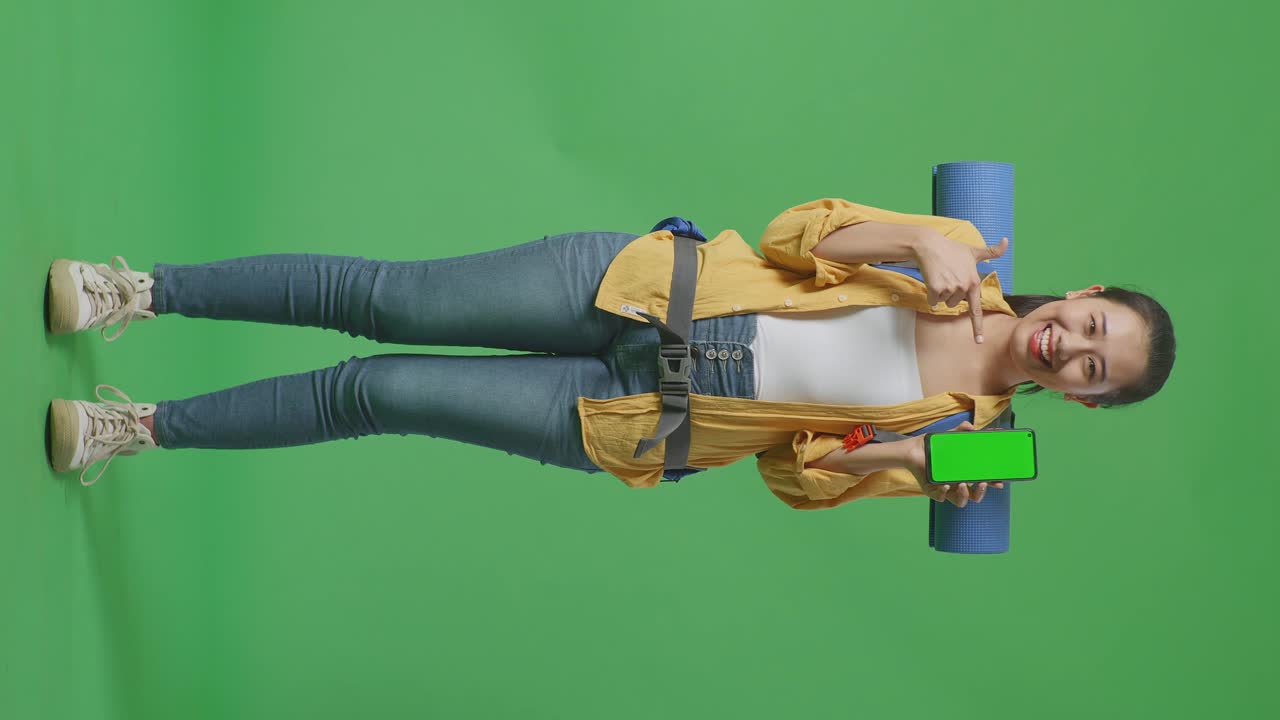 cuerpo lleno de excursionista femenina asiática con mountaineering mochila sonriendo y señalando a la imitación de pantalla verde teléfono inteligente mientras estaba de pie en el fondo de la pantalla verde en el estudio