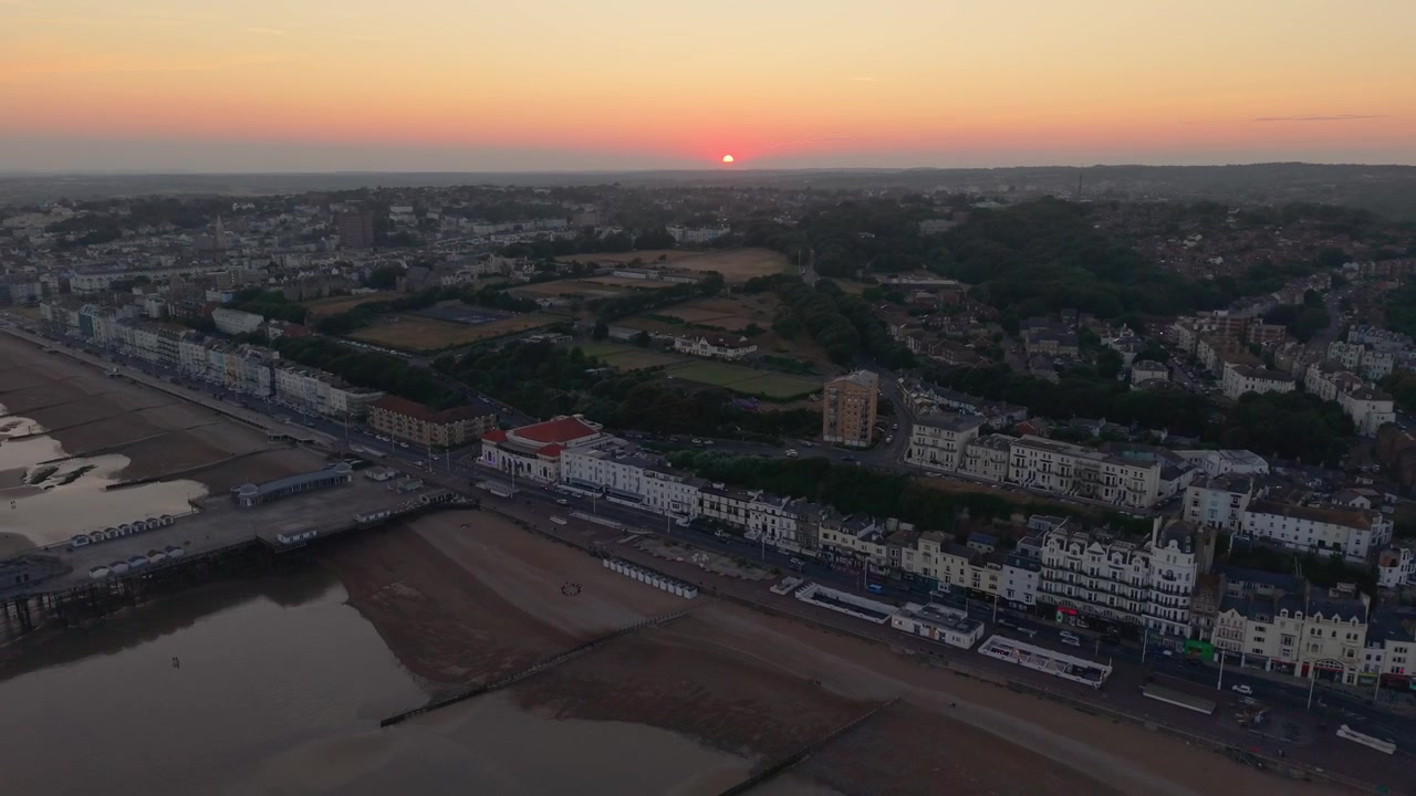 Drone si allontana in retromarcia dal molo di Hastings al tramonto, rivelando il lungomare, i campi sportivi e i tetti della città nell'East Sussex, Inghilterra.