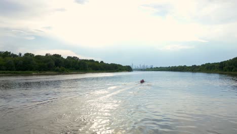 Canoe-rowing,-riverbank-vegetation,-water,-distant-city-silhouettes,-sky