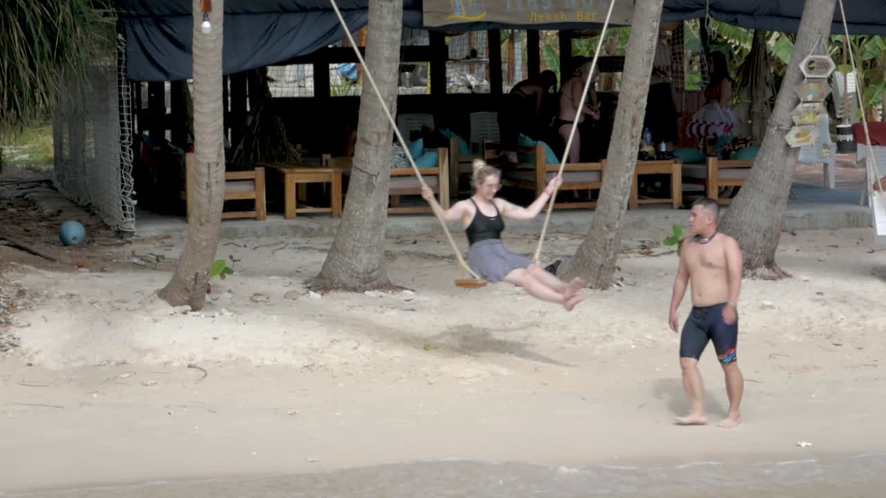 Woman swings on a rope swing at a tropical beachside bar with friends nearby