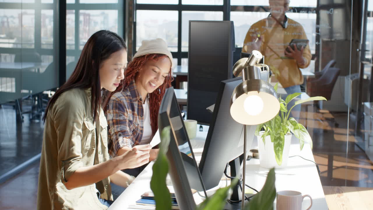 Diverse female team collaborating on coding project in modern office, using computers and tablets