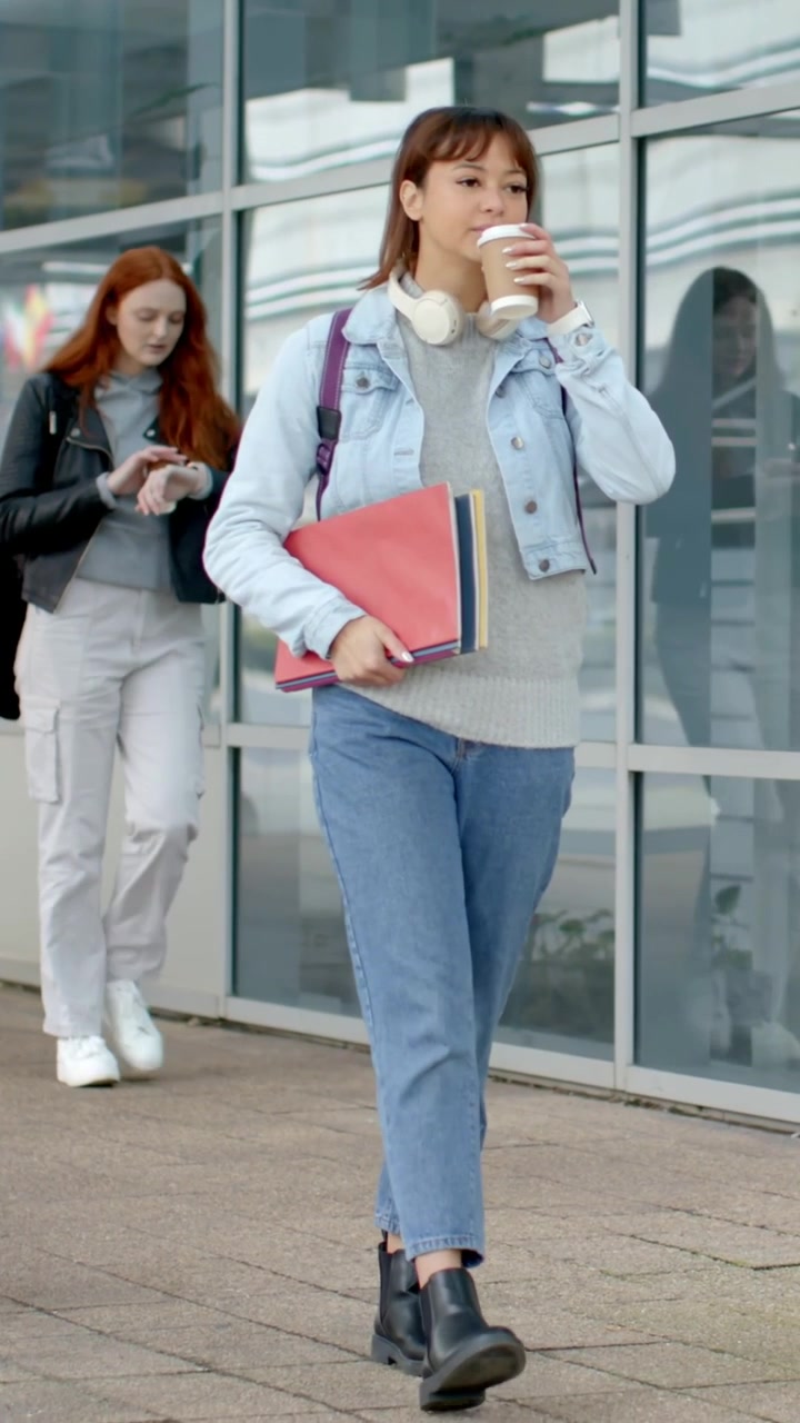 Vertikal video: Student i dongeri kommer mot kamera fra glassfasade, holder mapper og nipper til kaffe