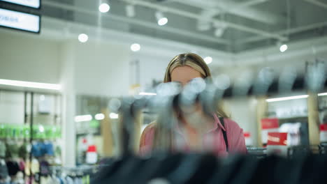 lady in pink shirt walks thoughtfully through clothes rack in shopping mall with black handbag, she carefully examines clothing in well-lit store as she stretch her hand to get one cloth