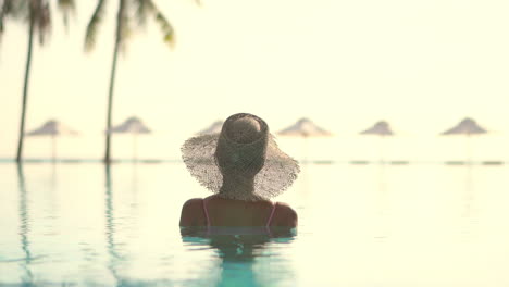 Rear-view-of-a-female-tourist-wearing-a-sun-hat,-sitting-half-submerged-in-pool-of-some-luxurious-resort,-looking-at-bright-sky-in-the-distance