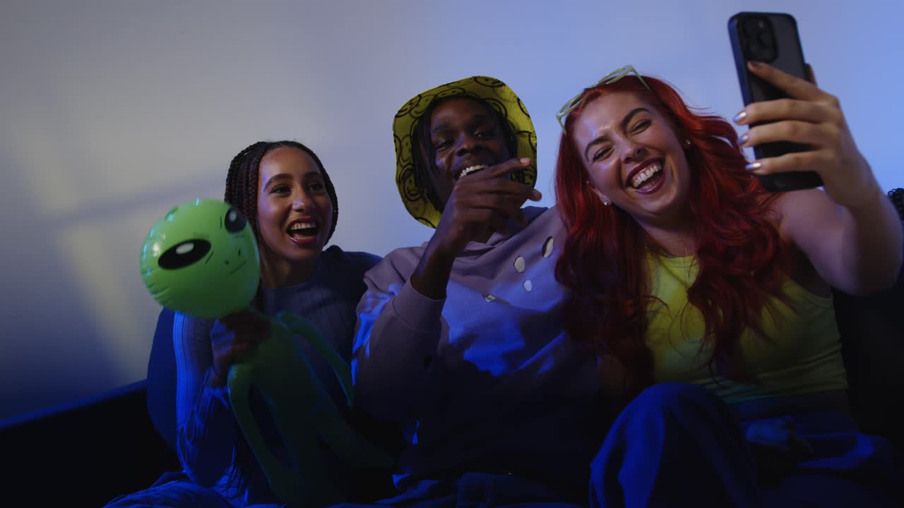 Studio Shot Of Group Of Gen Z Friends Sitting On Sofa Posing For Selfie On Mobile Phone At Night With Flashing Light 1