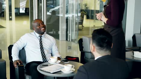 exitoso hombre de negocios afroamericano en ropa formal sonriendo y discutiendo el informe financiero con su colega caucásico en el café. joven camarera trayendo taza de café