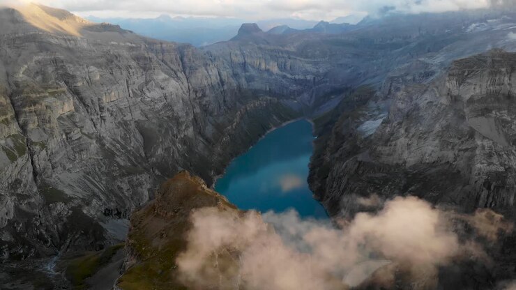 A flyover above lake Limmernsee in Glarus, Switzerland, with view of sunlit glowing clouds, a hydroelectric dam and reservoir surrounded by cliffs after sunset