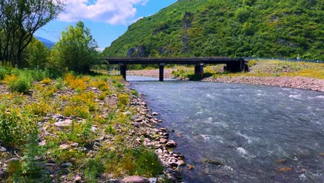 a serene view of a bridge over a flowing river surrounded by lush greenery and rocky shores under a bright blue sky