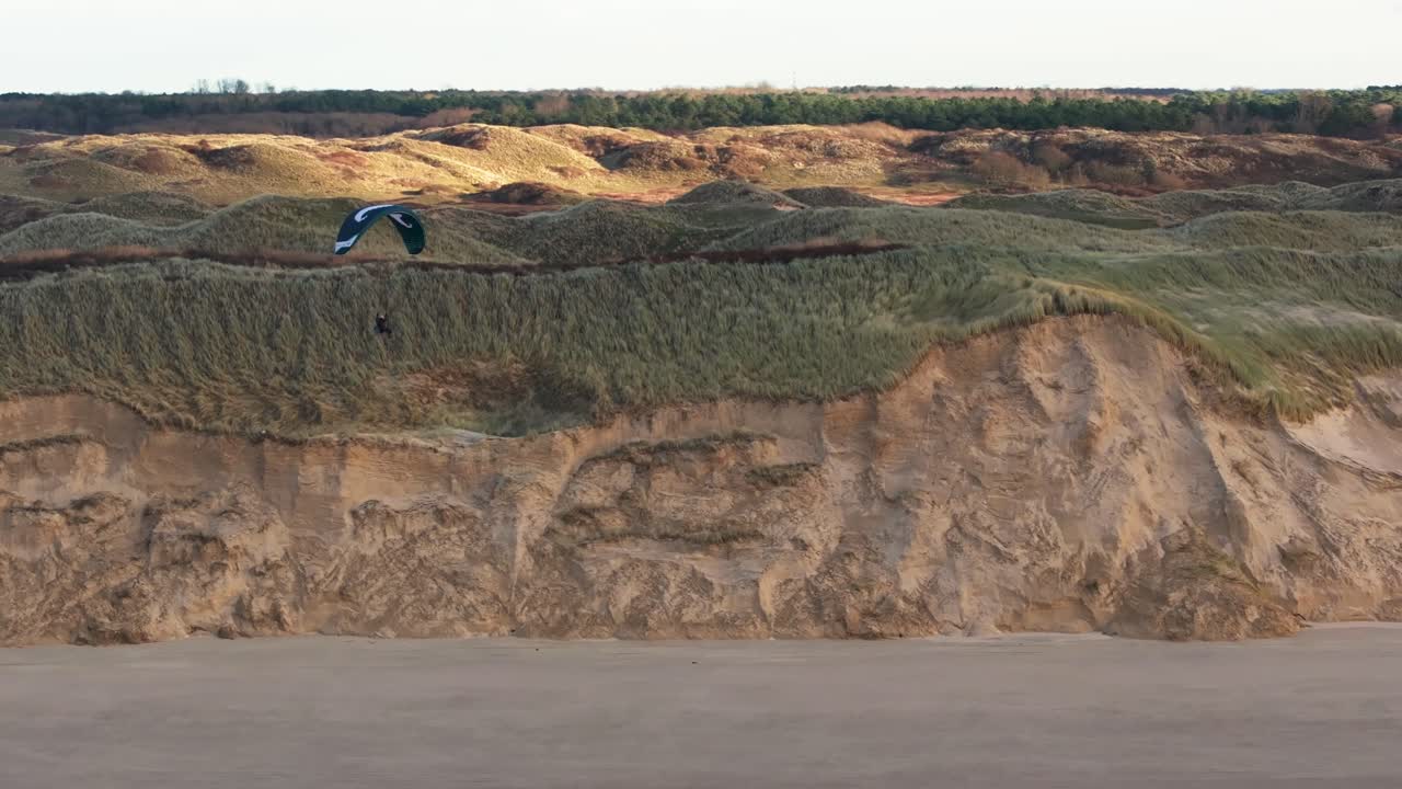 vista aérea baja de un parapente navegando a lo largo de las dunas de la costa de castricum en los países bajos con grandes dunas de arena y vegetación verde que cubre las dunas en la distancia