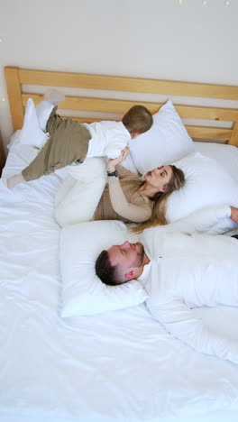 blonde woman lying on bed puts her kittle baby on her bent legs. mother plays with her son. bearded man lies beside. top view. vertical video.