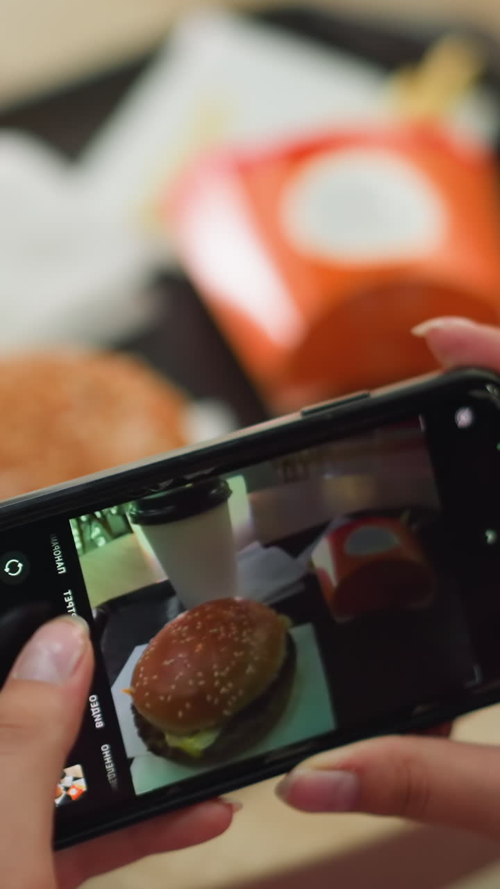 primer plano de una mujer tomando una foto de su comida con su teléfono inteligente, capturando una hamburguesa, papas fritas y café en una bandeja, la pantalla de su teléfono está ligeramente agrietada, mostrando la configuración de la comida
