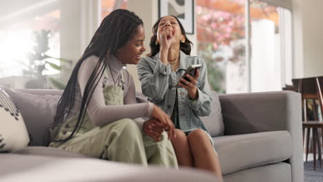 two friends laughing together while using a phone on the couch