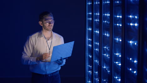 Male-network-engineer-doing-a-system-check-standing-in-the-server-room-with-his-laptop.-At-data-center-men-server-specialists-inspecting-working-system-and-hardware-of-rack-server-computer-cabinets