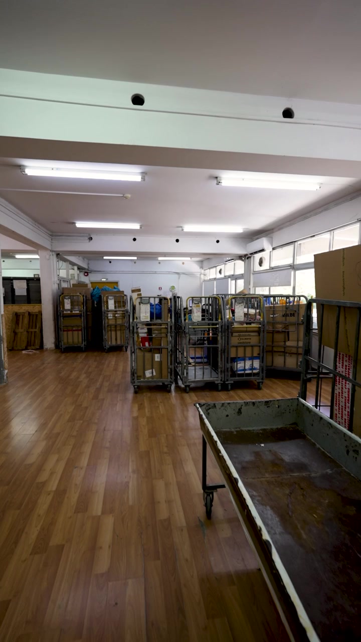 Caixas e prateleiras estão dispostas em uma área de armazenamento aberta. A luz vem de lâmpadas suspensas, e o espaço permanece livre de pessoas e itens.