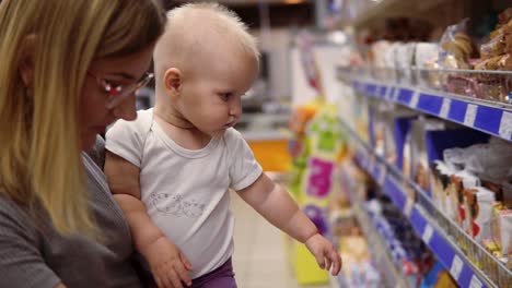 Young-mother-in-glasses-holding-her-child-in-her-arms-while-choosing-cookies-on-the-shelves-in-the-supermarket.-Thoughtful-mom