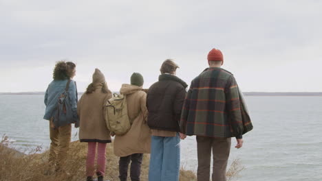 rear view of group of teenage friends in winter clothes holding hands and raising arms on top of the mountain on the beach on a windy day