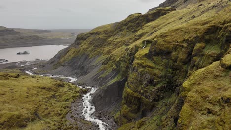 drone footage of a winding river cutting through moss-covered cliffs near sólheimajökull, iceland, with a distant glacial lake in view.