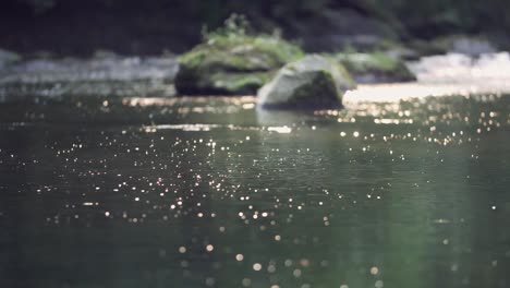 Slow-motion-of-creek-water-sparkling-from-sunlight,-mossy-rocks-behind