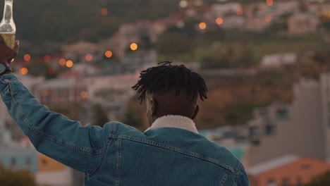 young-african-american-man-drinking-alcohol-with-arm-raised-enjoying-freedom-on-rooftop-making-toast-looking-at-city-skyline-sunset