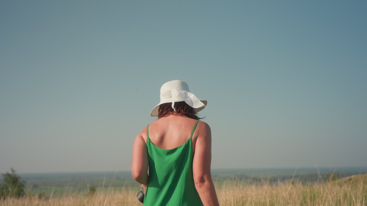 Side view of confident woman wearing green dress and white sunhat walking through dry golden grass field under clear blue sky holding camera, surrounded by calm nature and distant horizon