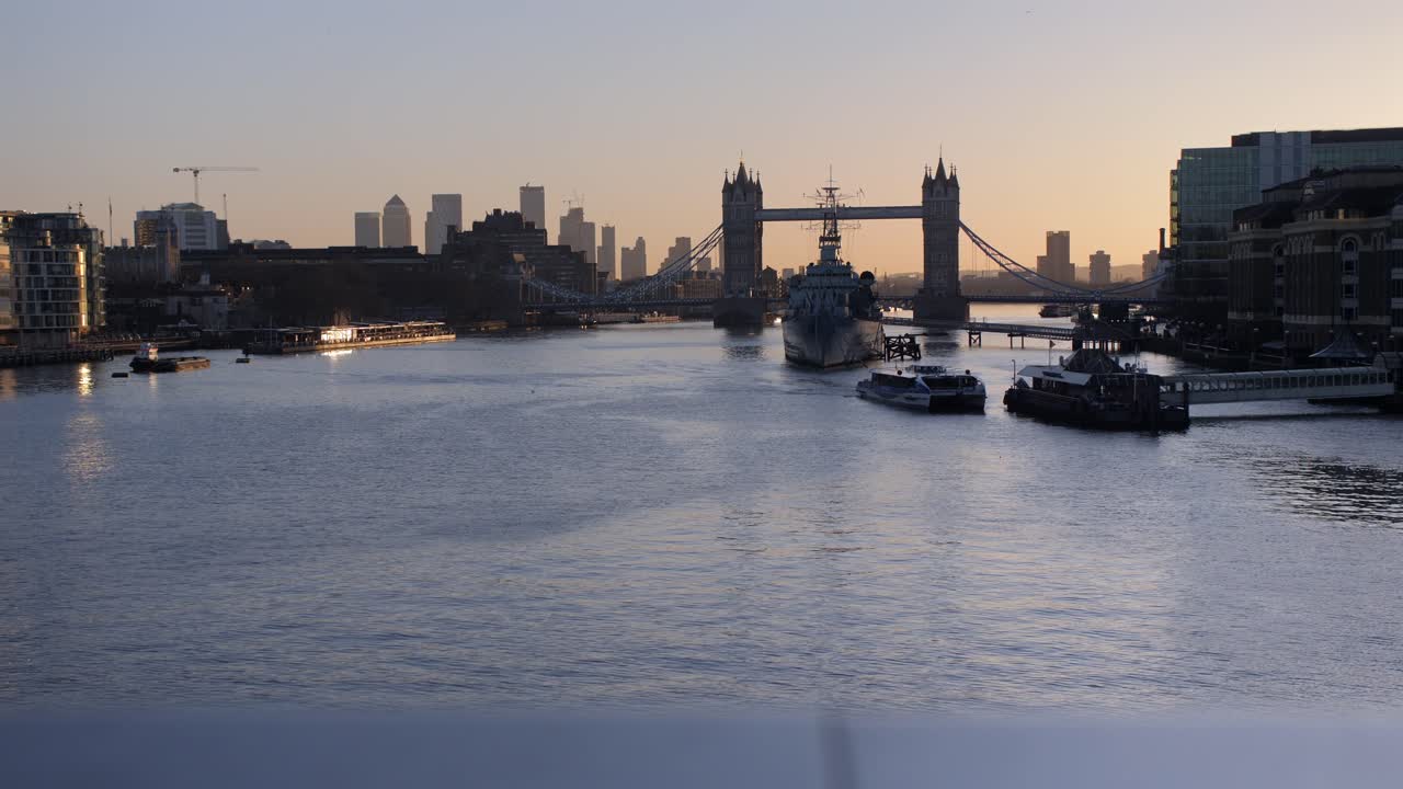 런던의 타워 브릿지와 해가 지는 테임즈 강의 넓은, 배와 따뜻한 저녁 빛을 반사하는 평온한 스카이라인.