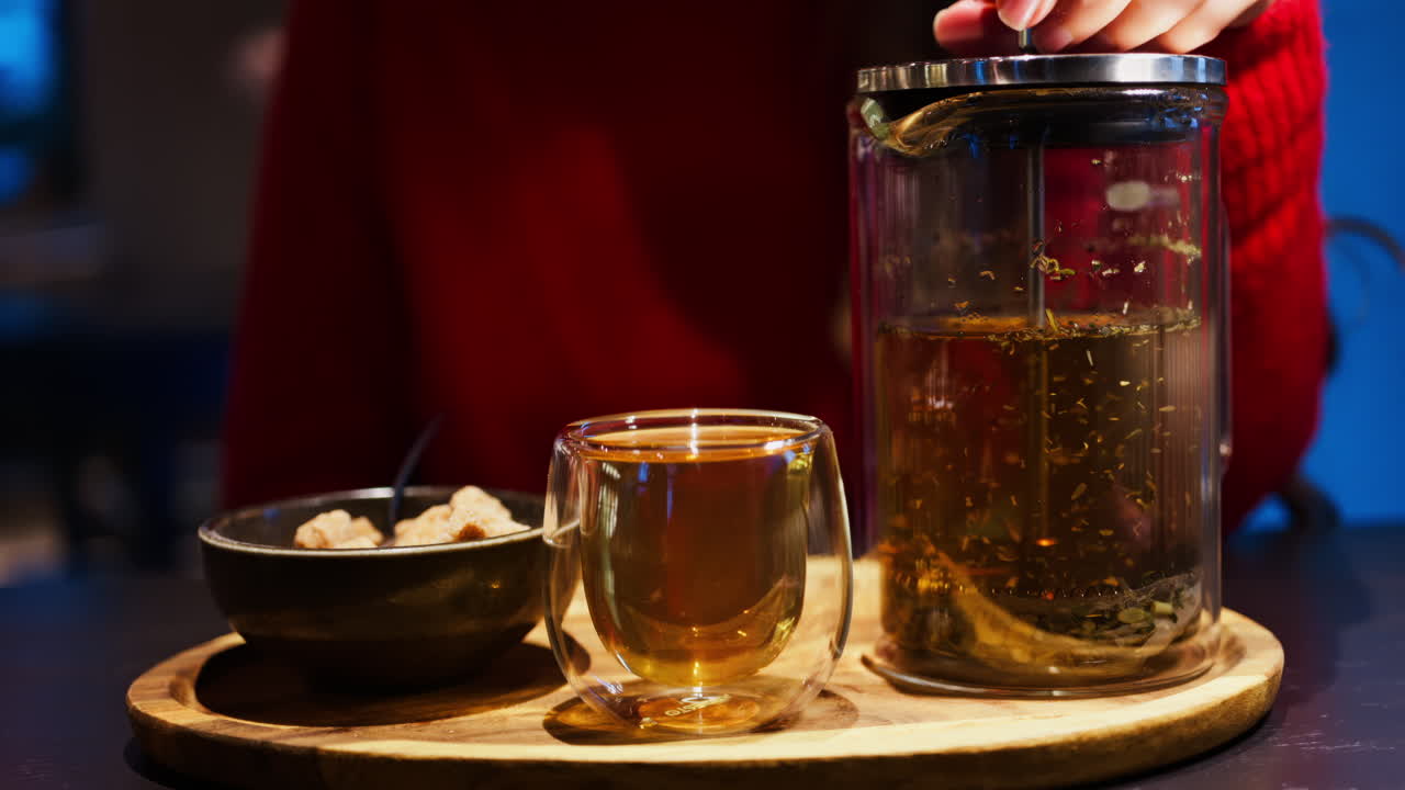 Close up of a woman pressing down the tea leaves with the help of a French press near a cup of tea at a restaurant