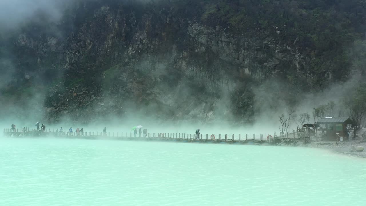 gente caminando sobre un puente con vapor saliendo del lago de azufre y la montaña detrás, aéreo