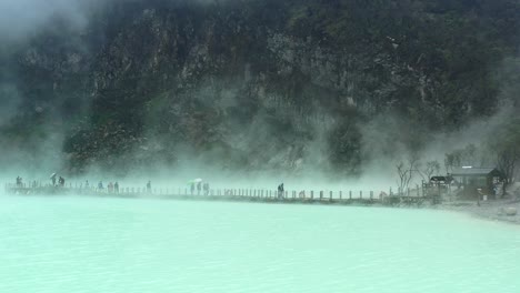 people walking on a bridge with steam rising from sulfur lake and mountain behind, aerial