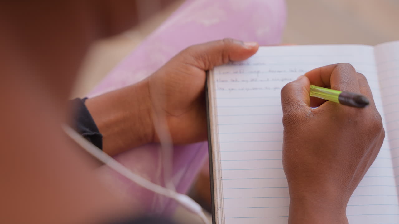 student writing in lined notebook closeup of hands holding green pencil, visible earphone cable and soft pink fabric backdrop, focused study and neat handwriting capturing reflective planning
