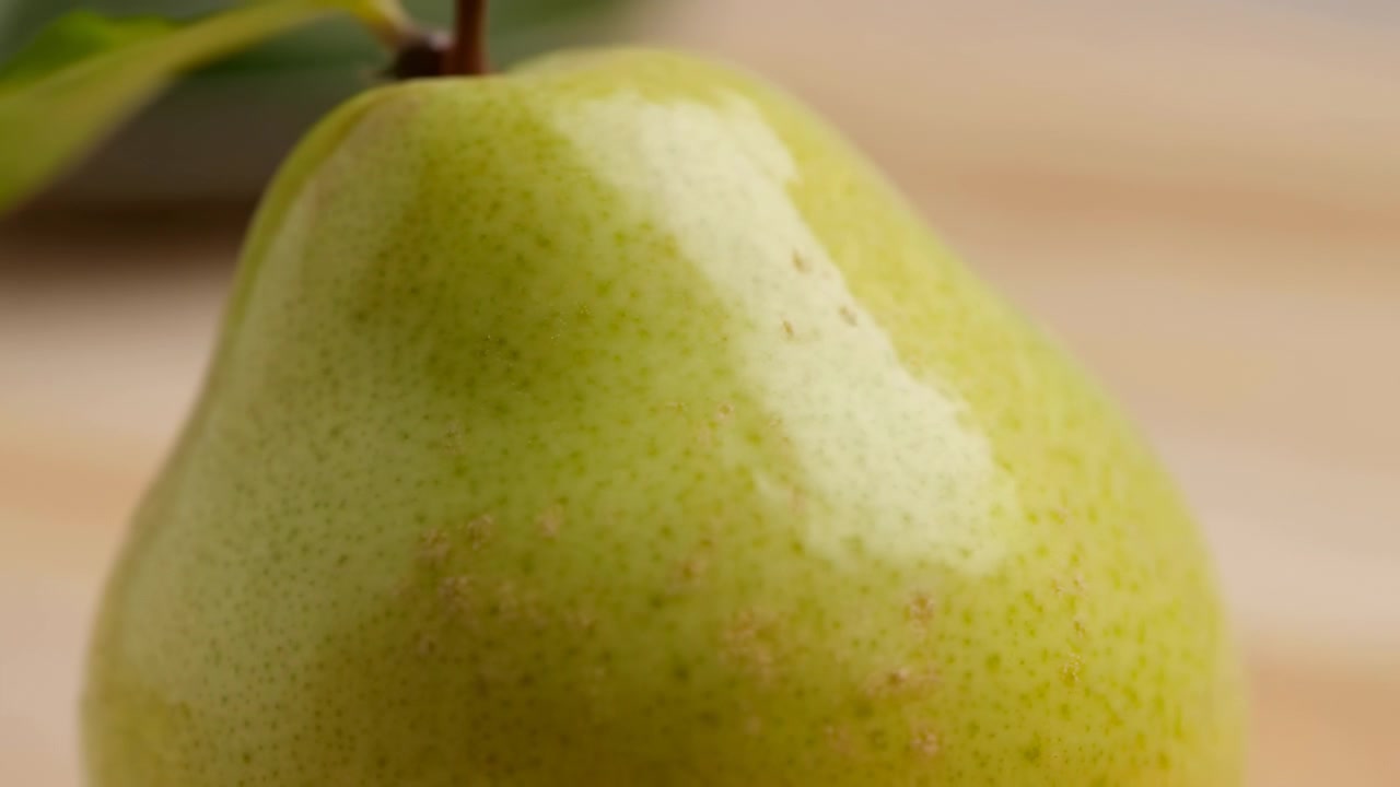 Una pera verde matura riempie l'inquadratura in primo piano, mostrando una superficie liscia e lucida, piccole macchie e un colore naturale fresco. Lo sfondo caldo e leggermente sfocato