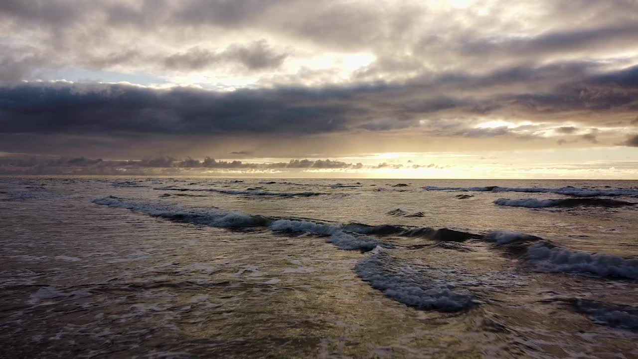 발트해 연안의 일몰 해변 위로 날아가는 아름다운 공중 샷, 높은 대비, 흐린 날씨, 잔잔한 파도 위로 뒤로 움직이는 저고도 광각 드론 샷