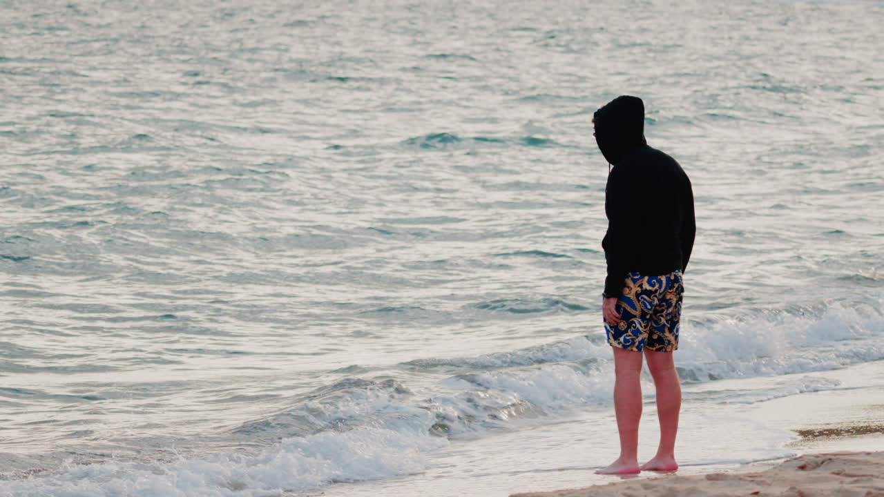 A man wearing a hoodie and patterned shorts stands barefoot by the shoreline, staring at the calm sea