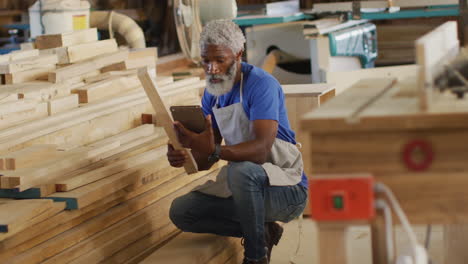 carpintero afroamericano con tableta digital eligiendo un tablón de madera en una carpintería
